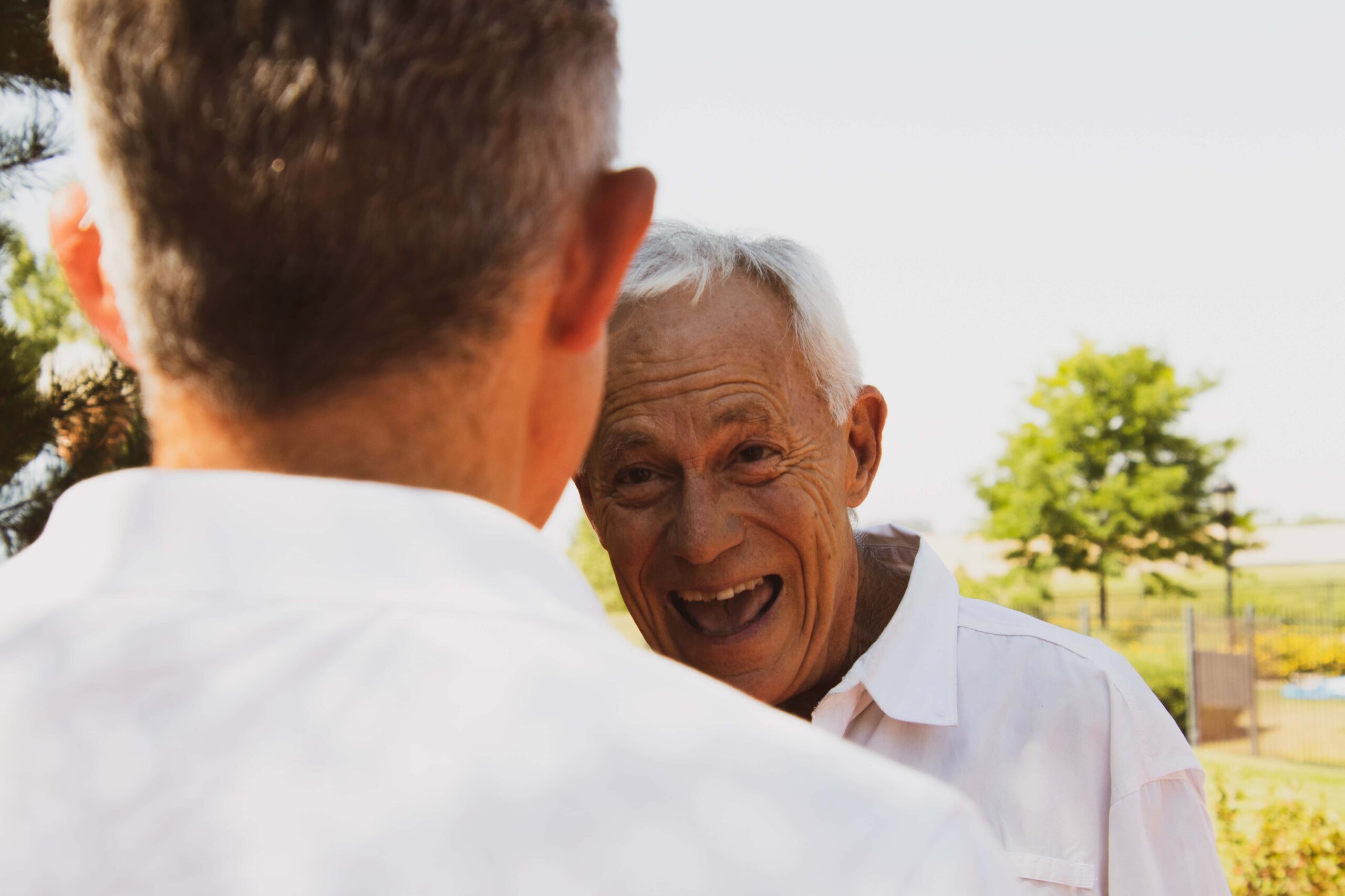Two older men in white shirts talk outdoors; one man faces the camera smiling while the other has his back to the camera. Trees and a fence are visible in the background.