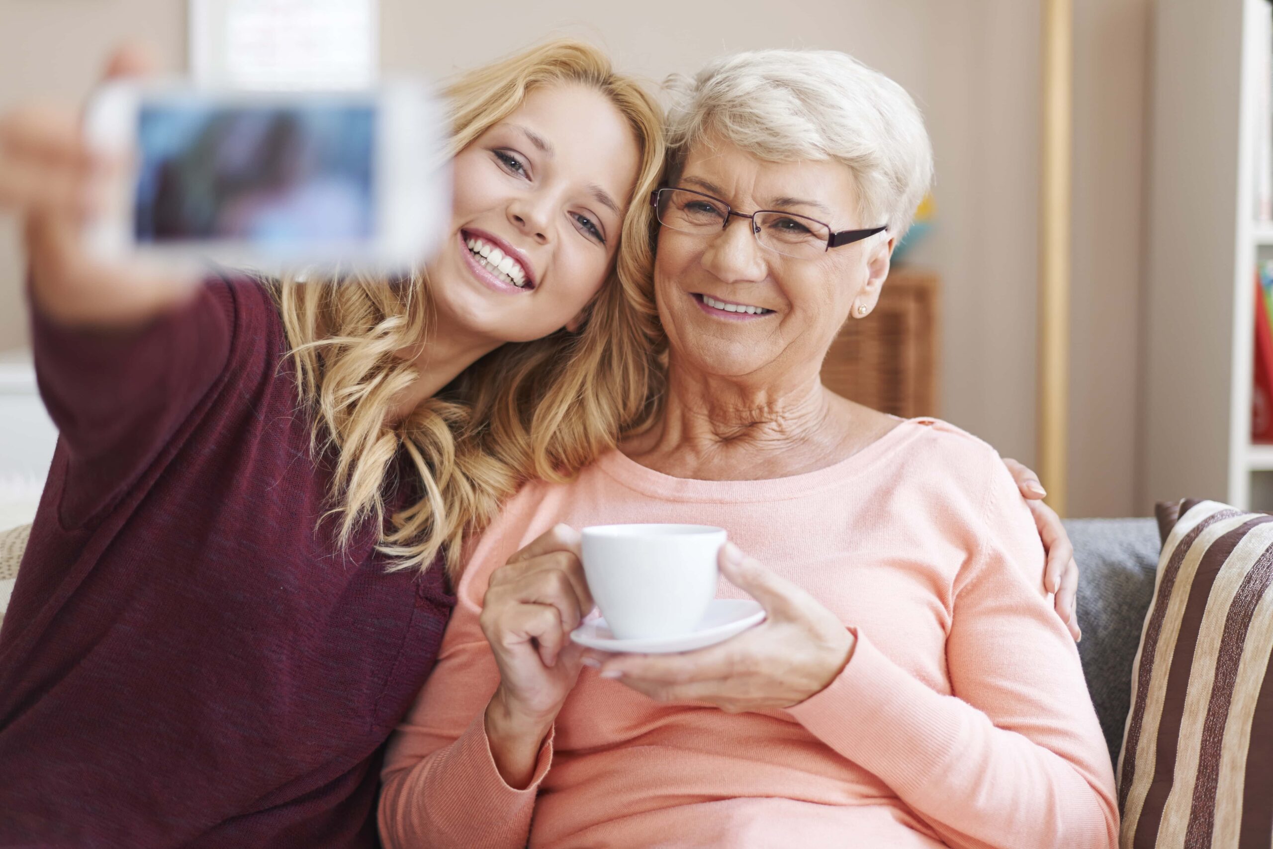 A young woman takes a selfie with an older woman who is holding a cup and smiling while sitting together on a couch.
