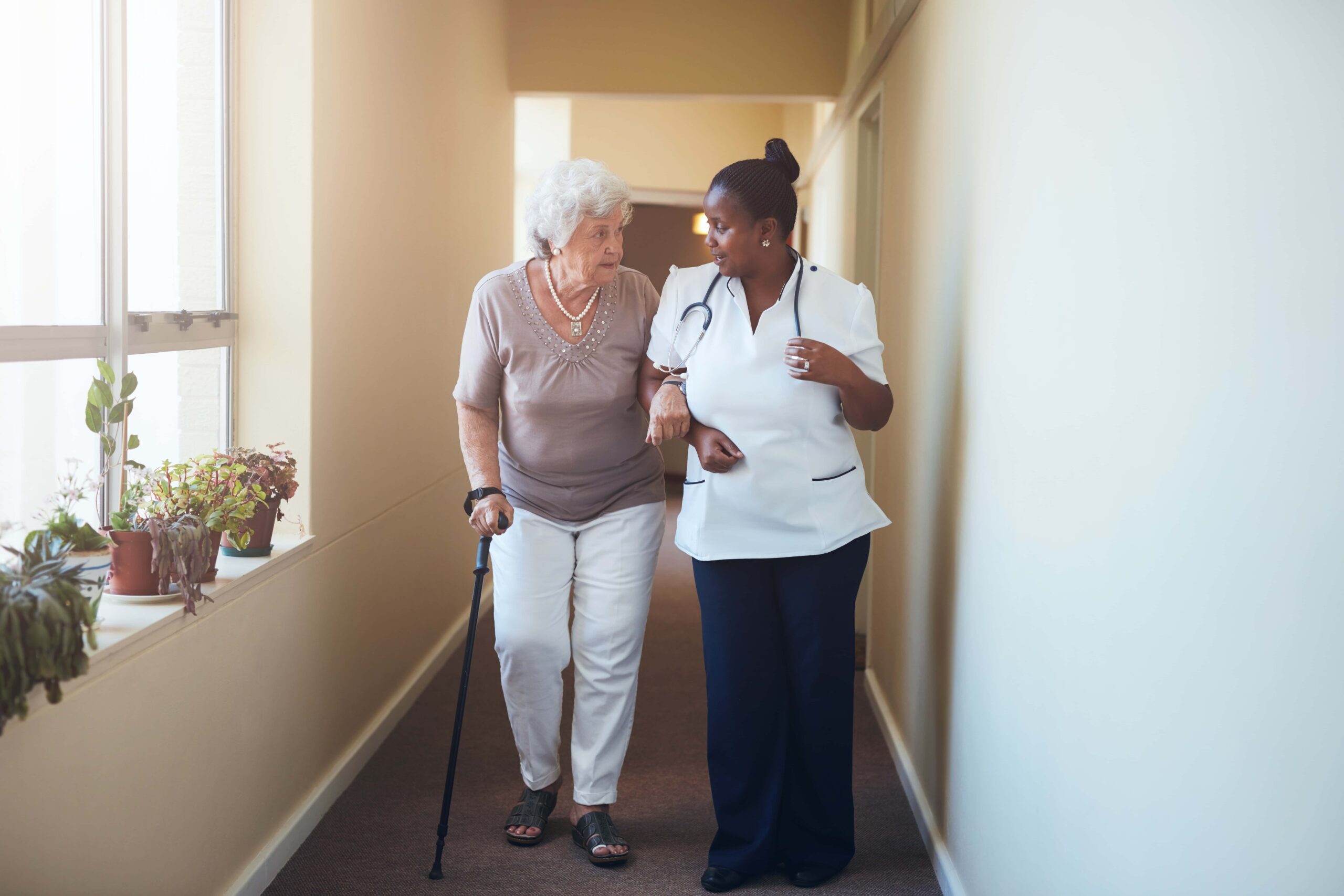 A nurse assists an elderly woman with a cane as they walk down a hallway together.