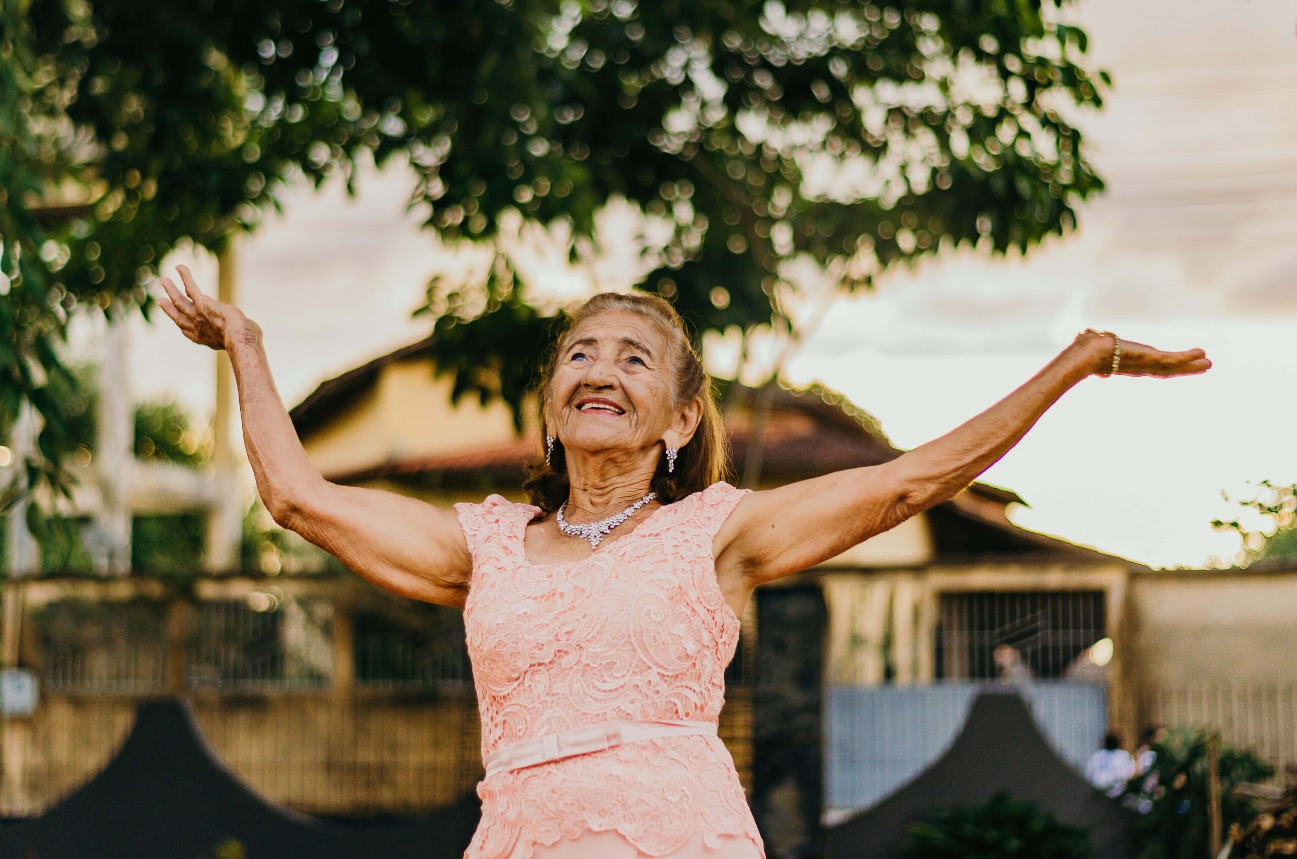 An elderly woman in a pink lace dress stands outdoors with arms raised, smiling, with trees and a house in the background.