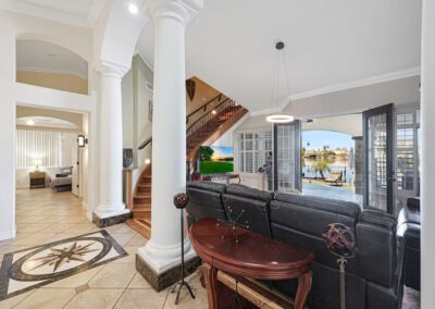 Spacious living room with black sectional sofa, round wooden table, columns, tile flooring, and French doors opening to a waterfront view. Wooden staircase visible in background.