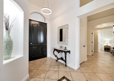 A foyer with a black front door, tiled floor, wall mirror above a decorative console table, and a large vase on display in a recessed wall niche.