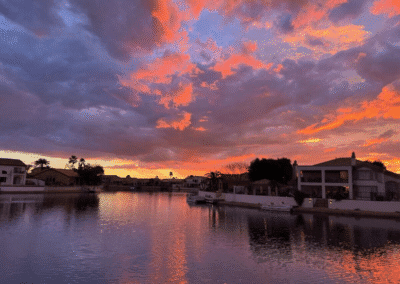 A residential waterfront neighborhood at sunset, with dramatic clouds reflecting orange and purple hues on the calm water.