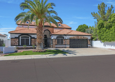 Two-story suburban house with a tile roof, black garage door, arched entryway, stone accents, and a large palm tree in the front yard.