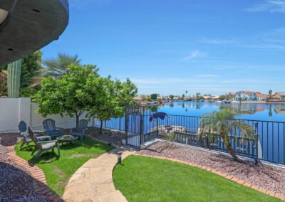 A fenced backyard with green grass, patio chairs, and a small tree overlooks a calm lake with houses along the shoreline under a clear blue sky.