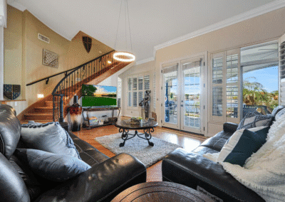 Modern living room with black leather sofas, a round coffee table, TV, and large windows facing a waterfront patio; a staircase leads upstairs.