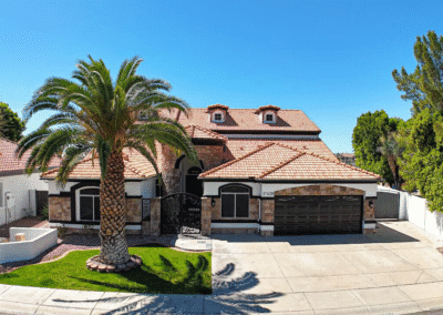 A two-story house with a red tile roof, stone accents, a large palm tree in front, and a double garage, set on a sunny day with clear blue sky.