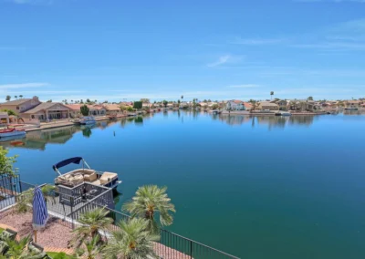 View of a calm, blue lake surrounded by houses, with a boat docked at a small pier and palm trees in the foreground under a clear sky.