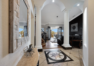A hallway with tiled floors and a compass rose design leads to a living area with columns, a wall clock, and large windows showing an outdoor view.